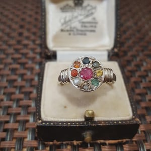 May include: A silver ring with a circular design, featuring a large, central red gemstone surrounded by smaller, colorful gemstones. The ring is displayed in a vintage jewelry box with a cream-colored interior.