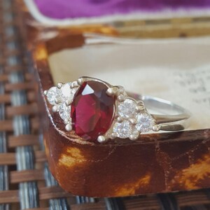 May include: A silver-toned ring featuring a large, oval-cut deep red gemstone at the centre, flanked by three small, clear, round-cut stones on each side. The band is smooth and metallic, showcasing a classic prong setting for all stones.