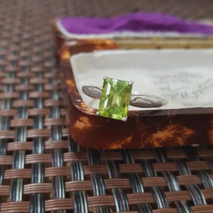 May include: A silver ring with a rectangular, light green gemstone. The ring is set against a backdrop of a vintage jewelry box with a brown exterior and a purple interior. The ring's band is thin and simple.