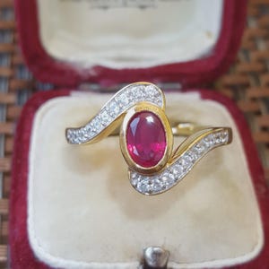 May include: A gold ring featuring a central oval-cut red ruby gemstone set in a bezel. The band has a bypass design with two curving arms, each channel-set with a row of small, clear, round-cut white stones that extend from the center setting.