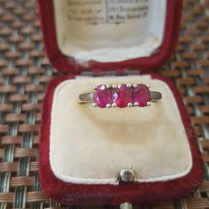 May include: A silver ring with three oval-cut, vibrant red gemstones. The ring is displayed inside a vintage red velvet jewelry box with a cream-colored interior, showcasing the ring's elegant design.