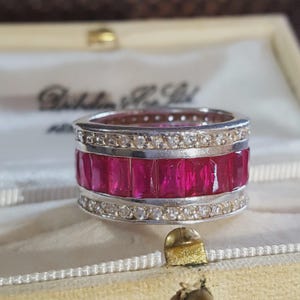 May include: A wide silver band ring featuring a central row of rectangular, vibrant red gemstones flanked by two rows of small, clear, round-cut white stones. The ring is constructed with a polished metal finish and a channel-set design.