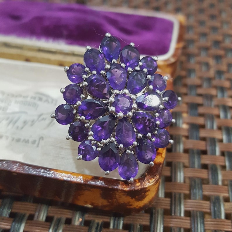 K&ouml;nnte beinhalten: Ein silberner Ring mit einem floralen Design, besetzt mit zahlreichen tiefvioletten Amethyst-Edelsteinen. Die Amethyststeine sind in verschiedenen Formen geschliffen und erzeugen einen funkelnden Effekt. Der Ring wird in einer Vintage-Schmuckschatulle pr&auml;sentiert.