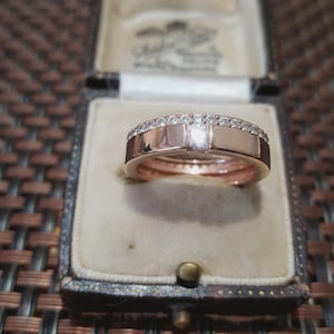 Può includere: Anello in oro rosa con una fascia di piccole pietre trasparenti e una pietra centrale più grande. L'anello è esposto in un portagioie vintage con interno color crema.