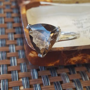 May include: A silver ring featuring a large, faceted, triangular-cut smoky quartz gemstone. The stone is held in place by three small prongs at each corner of the triangle, set upon a smooth, polished silver band that tapers towards the back.