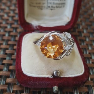 Peut inclure: Une bague en argent ornée d'une grosse pierre précieuse orange ronde. La bague présente un motif décoratif avec de plus petites pierres blanches et est présentée dans un écrin en velours rouge.