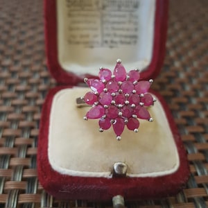 Può includere: Anello in argento con un motivo floreale, incastonato con numerose pietre preziose rosso rubino a forma di goccia. L'anello è presentato in un portagioie di velluto rosso.