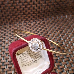 Peut inclure: Un bracelet jonc doré avec un motif circulaire central. Le cercle est orné de petites pierres précieuses transparentes entourant une pierre plus grande et transparente. Le bracelet est posé sur un écrin en velours rouge.