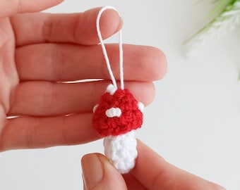 Può includere: Un piccolo portachiavi a uncinetto a forma di fungo con un cappello rosso e macchie bianche, e uno stelo bianco. Il portachiavi è attaccato a uno stringa bianca.
