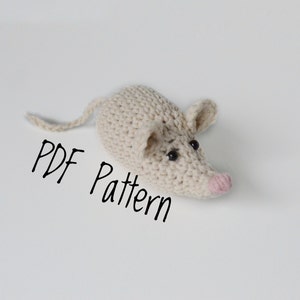 May include: A crocheted mouse amigurumi toy with black eyes and a pink nose. The toy is laying on a white surface with the text "PDF Pattern" written below it.