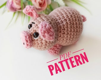 May include: A crocheted pink pig toy with black eyes. The pig is sitting on a white surface with a bouquet of pink flowers in the background. The text "PDF PATTERN" is written in pink on the white surface.