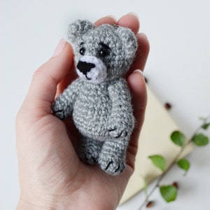May include: A small gray crocheted teddy bear keychain with black eyes and a white nose. The bear is being held in a person's hand.