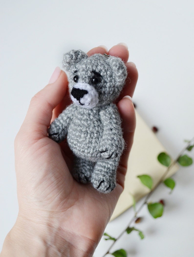 May include: A small gray crocheted teddy bear keychain with black eyes and a white nose. The bear is being held in a person's hand.