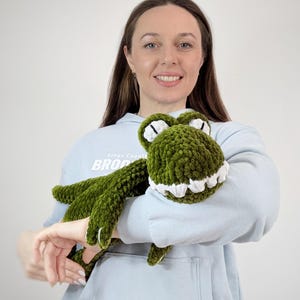 Puede incluir: Un peluche de cocodrilo verde hecho a mano con ganchillo, con dientes y ojos blancos. El peluche lo sostiene una persona que lleva una sudadera azul claro. El cocodrilo tiene un aspecto texturizado y difuso.