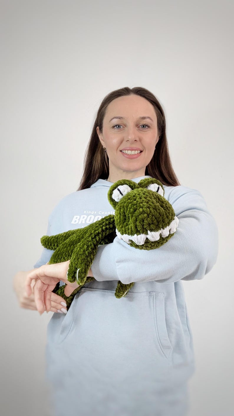 Puede incluir: Un peluche de cocodrilo verde hecho a mano con ganchillo, con dientes y ojos blancos. El peluche lo sostiene una persona que lleva una sudadera azul claro. El cocodrilo tiene un aspecto texturizado y difuso.