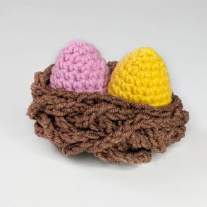 May include: A handcrafted crochet nest containing two eggs. One egg is pale pink, the other yellow. The nest is brown with a textured, woven look. The eggs are smooth and rounded, contrasting with the nest's rough texture. A charming Easter or spring decoration.