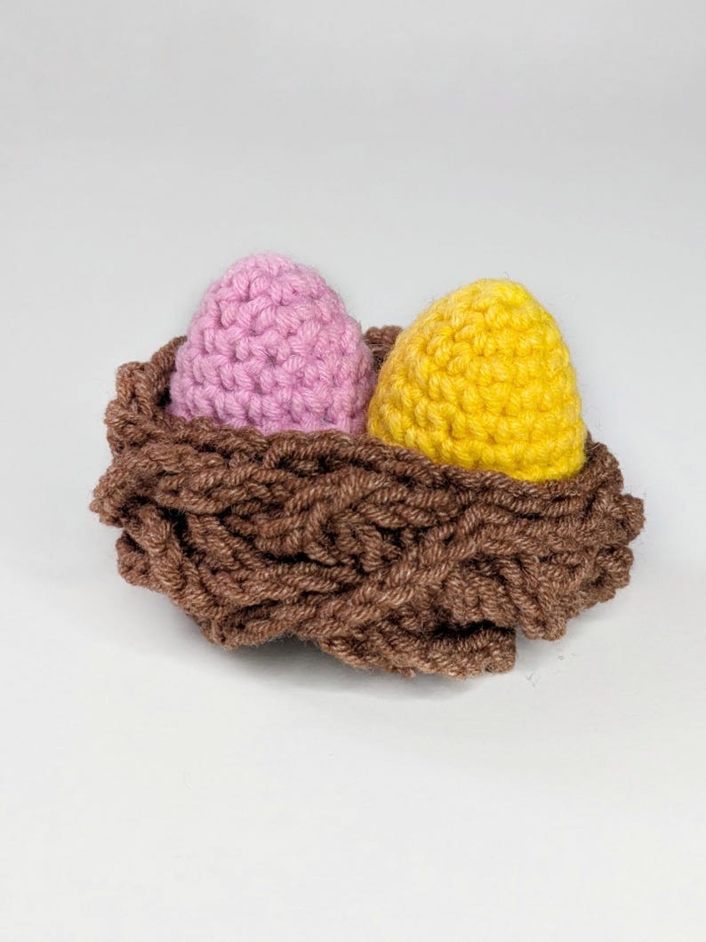 May include: A handcrafted crochet nest containing two eggs. One egg is pale pink, the other yellow. The nest is brown with a textured, woven look. The eggs are smooth and rounded, contrasting with the nest's rough texture. A charming Easter or spring decoration.