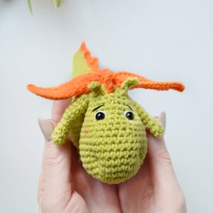 May include: A green crocheted dragon toy with orange wings. The dragon has black eyes and a cute expression.