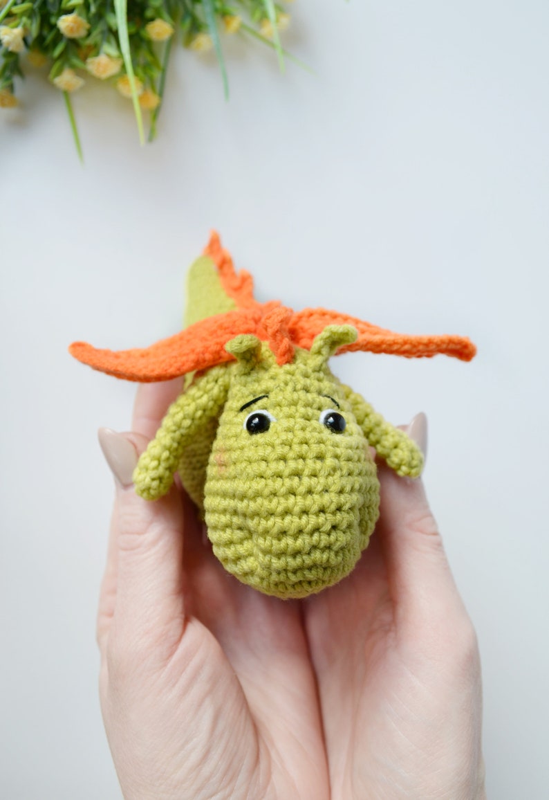 May include: A green crocheted dragon toy with orange wings. The dragon has black eyes and a cute expression.