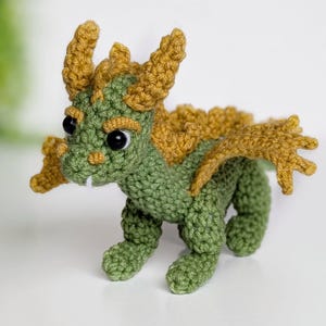 May include: A green crocheted dragon with yellow horns and wings. The dragon has black eyes and a toothy grin.
