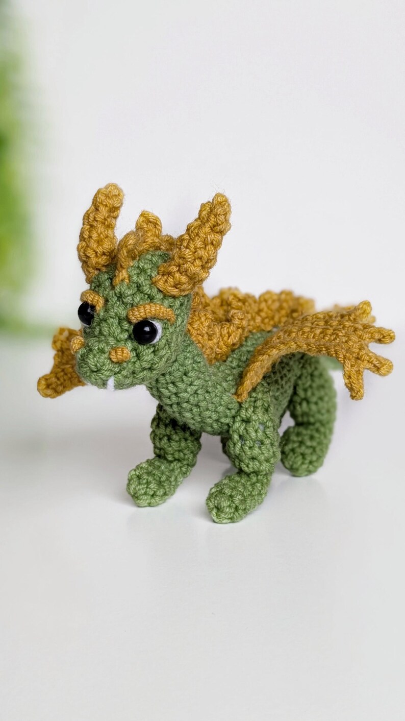 May include: A green crocheted dragon with yellow horns and wings. The dragon has black eyes and a toothy grin.