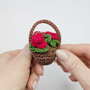 May include: A miniature crochet basket filled with red roses and green leaves. The basket is brown with a handle, and the flowers are a vibrant red. The handmade floral arrangement is held in a person's hands.