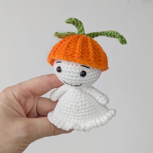 May include: A small, handmade crochet doll featuring a white, rounded body with a flared base and a bright orange, ribbed hat-like top. The doll has two black bead eyes and a simple stitched mouth, topped with two green, leaf-like crochet stems.
