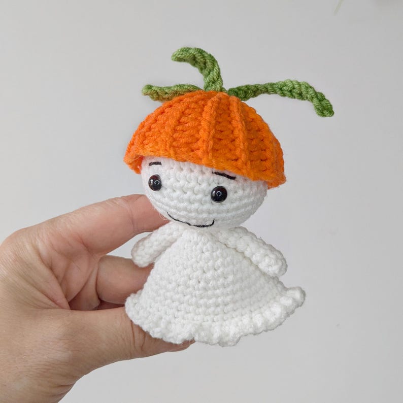 May include: A small, handmade crochet doll featuring a white, rounded body with a flared base and a bright orange, ribbed hat-like top. The doll has two black bead eyes and a simple stitched mouth, topped with two green, leaf-like crochet stems.