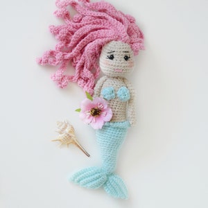 May include: A crocheted mermaid doll with pink hair, a blue tail, and a pink flower. The doll is holding a seashell.