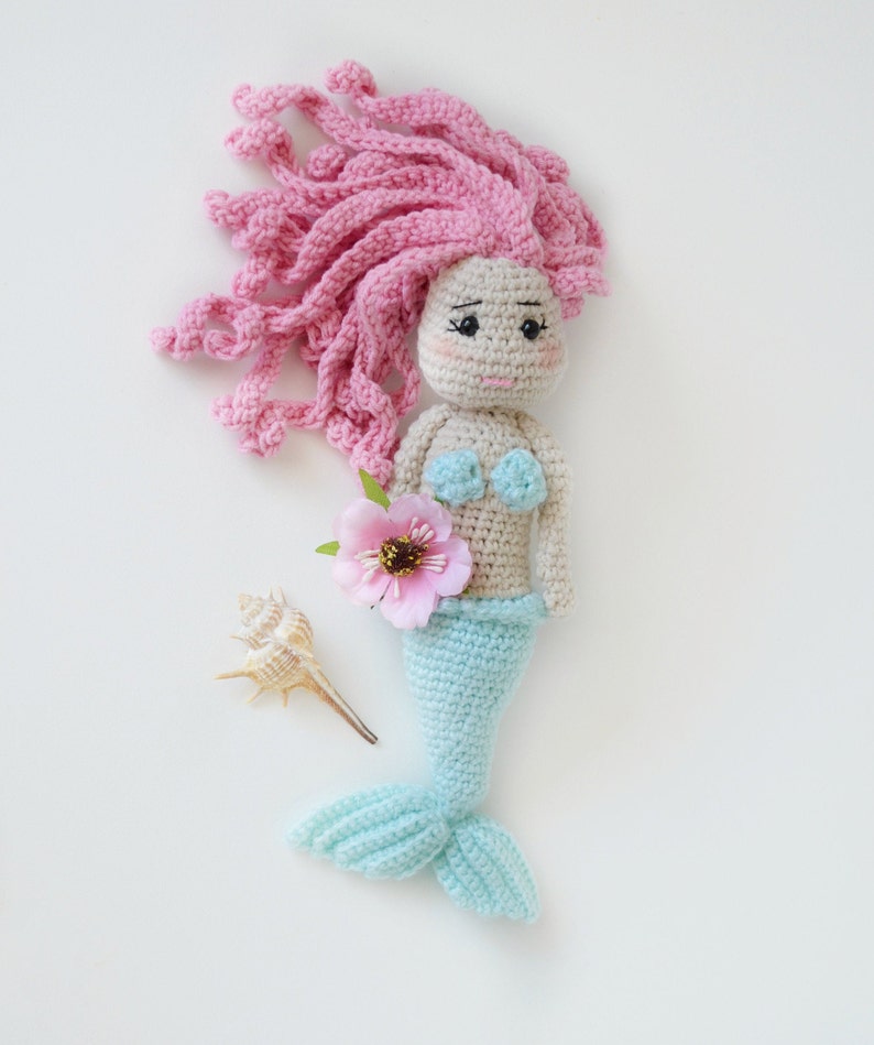 May include: A crocheted mermaid doll with pink hair, a blue tail, and a pink flower. The doll is holding a seashell.