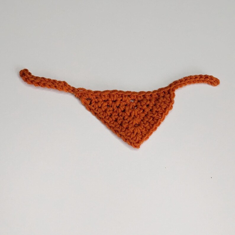 May include: A triangular-shaped bandana crafted from burnt orange yarn using a crochet technique. The accessory features a textured, knotted stitch pattern throughout the main body and includes two thin, extended ties on either side for fastening.
