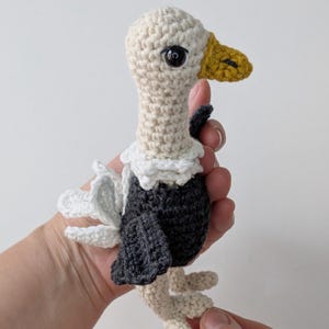 May include: A small crochet vulture toy featuring a cream-colored head and neck, a dark gray body, and white ruffled neck feathers. The bird has a textured mustard yellow beak, black plastic eyes, and white wing accents on its back.