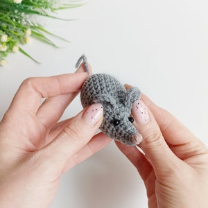 May include: A small gray crocheted mouse with black eyes and a pink nose. The mouse is being held by two hands.
