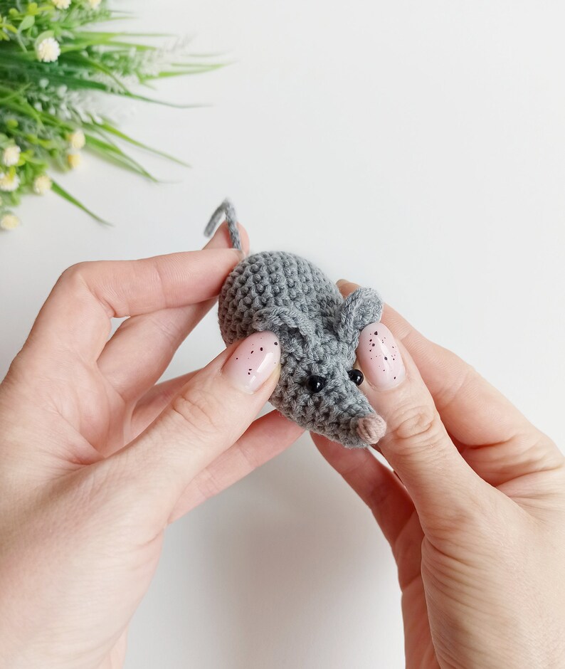 May include: A small gray crocheted mouse with black eyes and a pink nose. The mouse is being held by two hands.