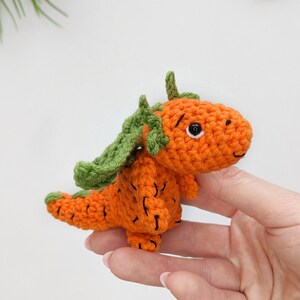 May include: A crocheted carrot-shaped dragon toy with green leaves for wings. The toy is orange and has black eyes.