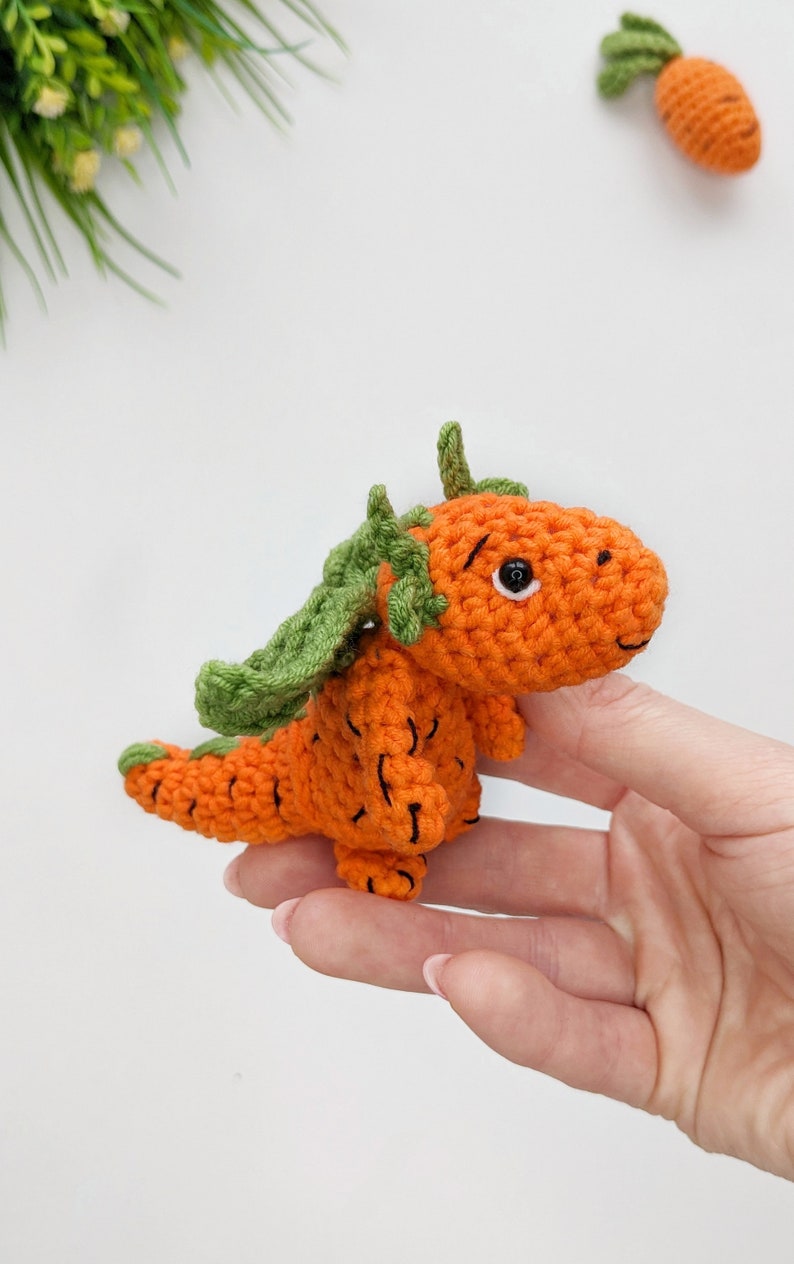 May include: A crocheted carrot-shaped dragon toy with green leaves for wings. The toy is orange and has black eyes.