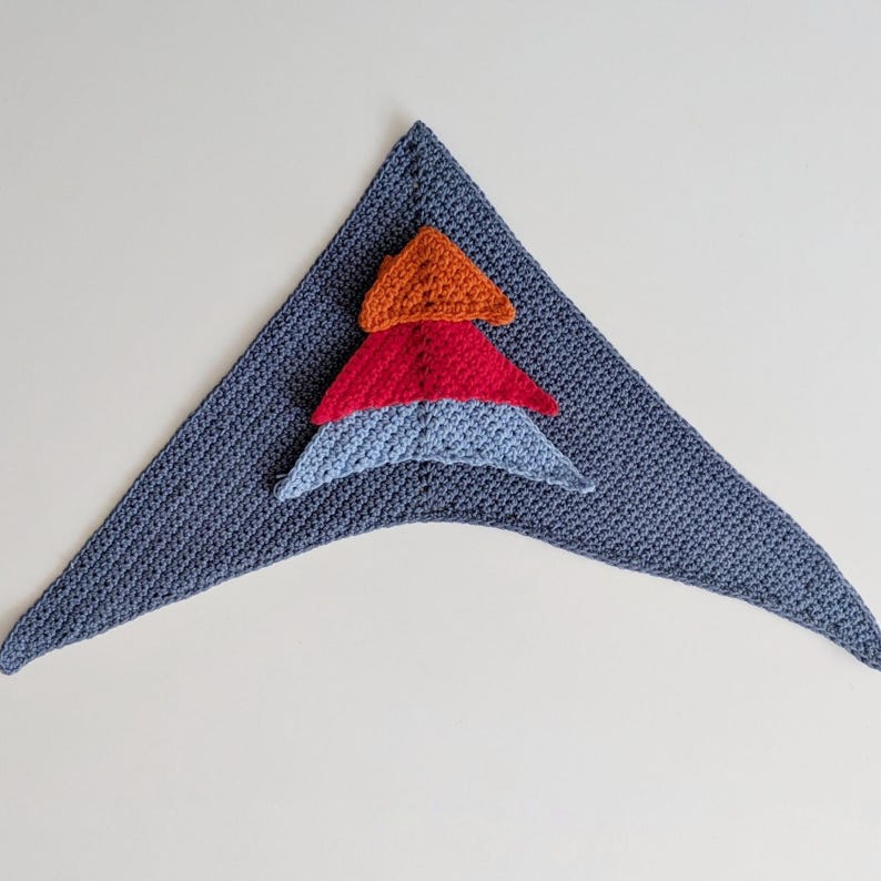 May include: A triangular crochet scarf in slate blue featuring a stacked applique design of three smaller triangles in orange, red, and light blue. The textile has a consistent knit texture throughout the entire piece, showcasing a layered geometric pattern.