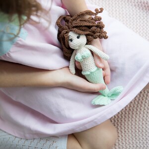 May include: A young person is holding a crocheted mermaid doll with brown hair, a green tail, and a white top. The doll is sitting in the person's lap.