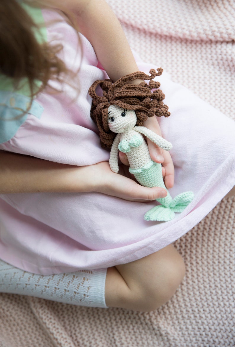 May include: A young person is holding a crocheted mermaid doll with brown hair, a green tail, and a white top. The doll is sitting in the person's lap.