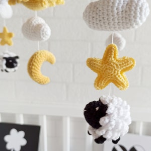May include: A white and yellow crocheted baby mobile with a cloud, stars, a crescent moon, and two sheep. The mobile is hanging from a white metal frame.