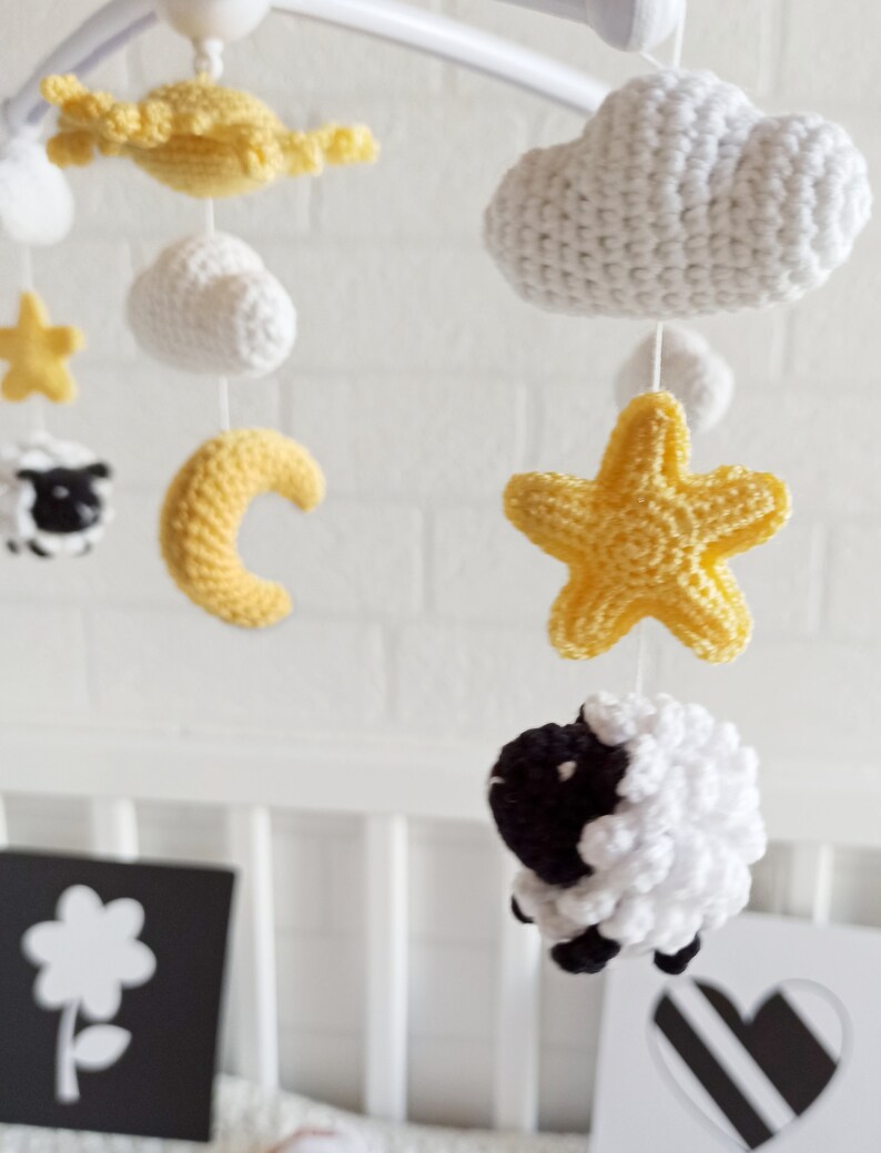 May include: A white and yellow crocheted baby mobile with a cloud, stars, a crescent moon, and two sheep. The mobile is hanging from a white metal frame.