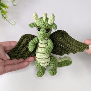 May include: A green crocheted dragon with light green stripes and black eyes. The dragon has wings that are spread out.