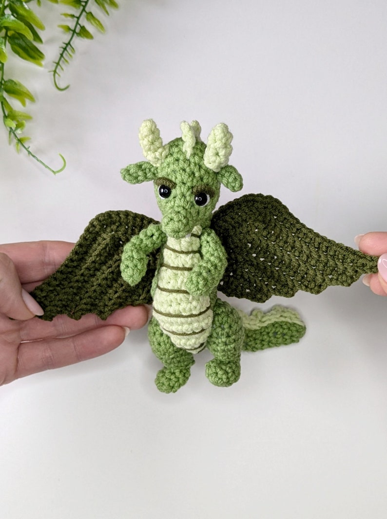 May include: A green crocheted dragon with light green stripes and black eyes. The dragon has wings that are spread out.