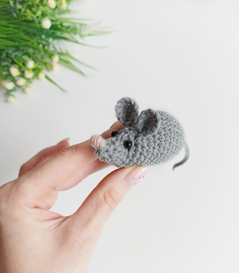 May include: A small gray crocheted mouse with black eyes and a pink nose. The mouse is being held by a hand.