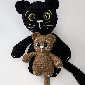 May include: A large black crocheted cat plush toy with glittery gold-rimmed eyes and white stitched whiskers, holding a smaller brown crocheted cat plush toy that also features white stitched whiskers and small white paw details on its feet.