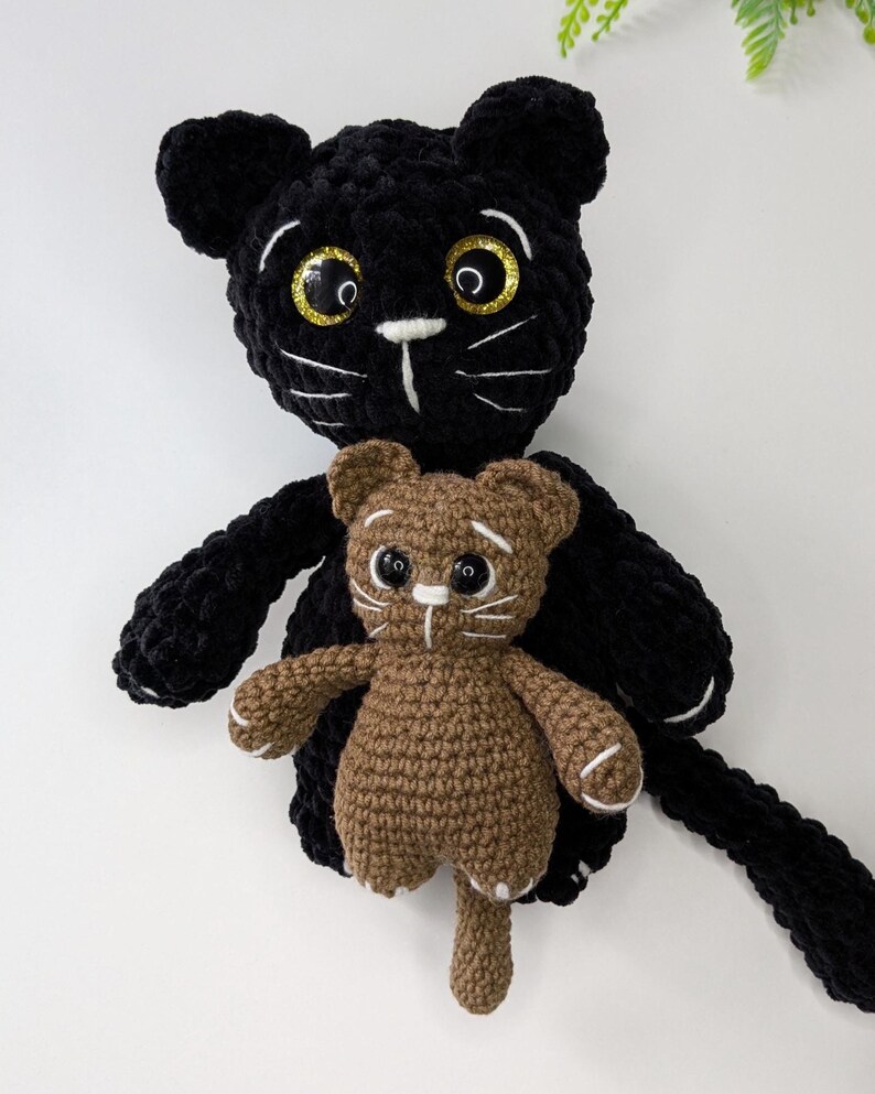 May include: A large black crocheted cat plush toy with glittery gold-rimmed eyes and white stitched whiskers, holding a smaller brown crocheted cat plush toy that also features white stitched whiskers and small white paw details on its feet.