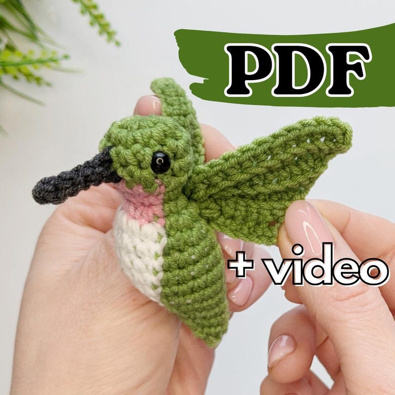 May include: A crocheted hummingbird amigurumi toy with green wings, a pink and white body, and black bead eyes. The toy is being held in a hand and the text "PDF + video" is visible in the image.