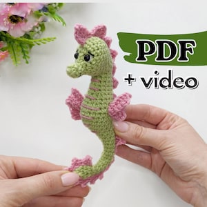Puede incluir: Un juguete de amigurumi de caballito de mar tejido a crochet con aletas rosas y cuerpo verde. El juguete está siendo sostenido en las manos de una persona. El texto "PDF + video" es visible en el fondo.