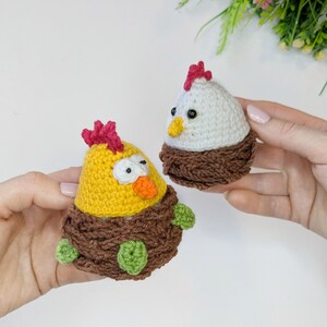 May include: Two handmade crochet chicken figurines. One is yellow with a red comb and orange beak, the other is white with a red comb and yellow beak. Both are set in brown crochet nests with green accents.
