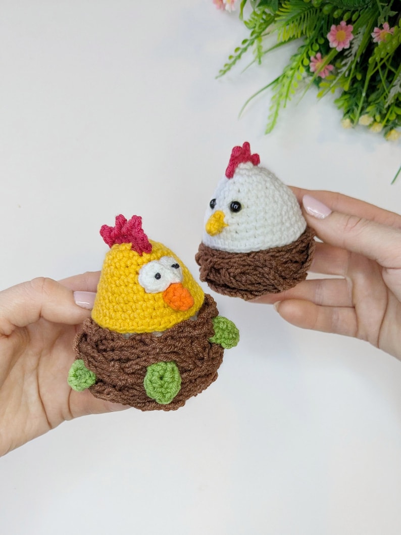May include: Two handmade crochet chicken figurines. One is yellow with a red comb and orange beak, the other is white with a red comb and yellow beak. Both are set in brown crochet nests with green accents.